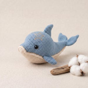 May include: A blue crocheted dolphin toy with a cream-colored underbelly and a small embroidered gold star on its forehead. The dolphin features black plastic safety eyes and a smooth, textured yarn finish, positioned next to a piece of driftwood.
