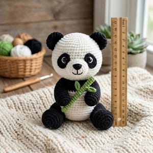 May include: A handmade crochet panda bear with black and white coloring, holding a green bamboo stalk. The panda is approximately 6 inches tall, sitting on a cream-colored knitted surface. A wooden ruler is next to the panda.