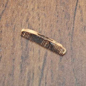 May include: A small, light brown comb with fine teeth. The comb is made of a light-colored material and has a decorative cutout design. The comb is lying on a wooden surface.