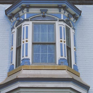 Peut inclure: Gros plan sur une baie vitrée avec un design peint en bleu et blanc. La fenêtre comporte plusieurs vitres et est encastrée dans un mur de briques bleu clair. La garniture présente des éléments décoratifs.