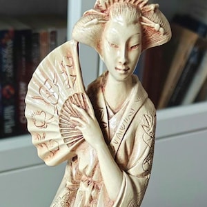 Puede incluir: Figurilla de color marfil de una geisha sosteniendo un abanico. La geisha lleva un kimono detallado y tiene un peinado elaborado. El abanico está abierto, revelando patrones intrincados. La estatua es un artículo decorativo, probablemente para exhibición.