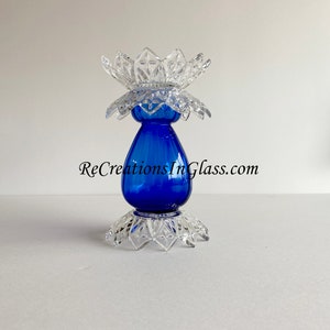 May include: A cobalt blue glass vase with a clear glass base and top. The base and top are shaped like a flower with multiple points. The vase is decorated with a clear glass design. The text "ReCreationsInGlass.com" is visible on the vase.