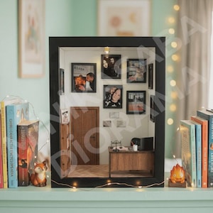 Könnte beinhalten: Ein Miniatur-Raum-Diorama in einem schwarzen rechteckigen Rahmen mit einem Schreibtisch aus Holz, einer geschlossenen Holztür und mehreren gerahmten Fotos an den weißen Wänden. Die Szene wird durch ein kleines Licht und Lichterketten beleuchtet.