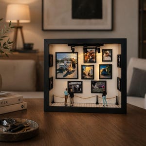 May include: A black rectangular shadow box frame designed as a miniature art gallery with internal lighting. The interior features multiple framed photographs on the back wall, a tiled floor, and three small figurines standing behind a rope barrier.