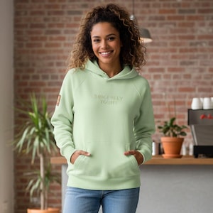 May include: A light green hoodie with the text "Sincerely Yours" embroidered on the front. The hoodie has a kangaroo pocket and a hood. The person in the image is wearing blue jeans. A small brown patch is on the left sleeve.