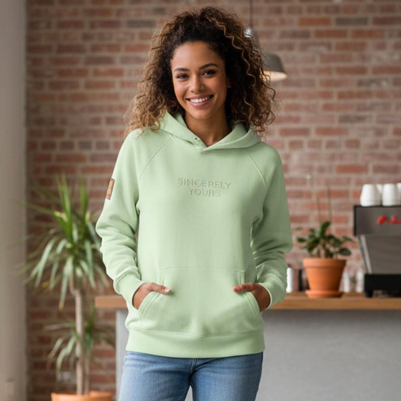 May include: A light green hoodie with the text "Sincerely Yours" embroidered on the front. The hoodie has a kangaroo pocket and a hood. The person in the image is wearing blue jeans. A small brown patch is on the left sleeve.