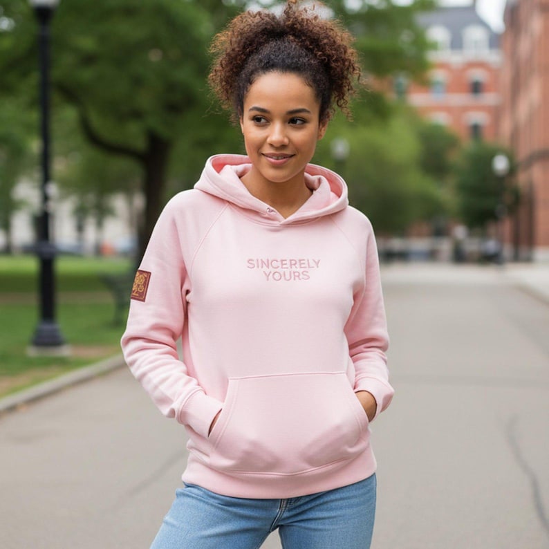 May include: A light pink hoodie with the text "Sincerely Yours" on the front. The hoodie has a kangaroo pocket and a hood. A small patch is on the left sleeve. The person is wearing blue jeans.