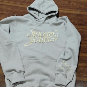 以下が含まれることがあります： フードとフロントポケットが付いた、明るいグレーのパーカー。「Sincerely Yours」の文字が胸にクリーム色で刺繍されています。左袖には星座のデザインが刺繍されています。