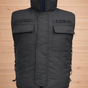 Könnte beinhalten: Eine dunkelgraue Steppweste mit hohem Kragen und mehreren Taschen. Die Weste hat zwei große Fronttaschen und kleinere Taschen auf der Brust. Sie wird auf einer Schaufensterpuppe vor einem Holzpaneel-Hintergrund präsentiert.