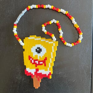 May include: A yellow beaded necklace featuring a SpongeBob SquarePants popsicle design. The necklace has a beaded chain with red, yellow, and white beads. The word "SUBTRONICS" is spelled out on the necklace.