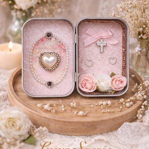 May include: Open, decorative tin box with a pink floral design. One side features a heart-shaped pearl pendant and the other a cross and pink roses. The box is on a wooden tray, surrounded by flowers and pearls. The text "Lueur Sacrée ATELIER" is visible.