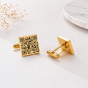 Op de afbeelding: Een paar goudkleurige manchetknopen. Elke manchetknop heeft een vierkante voorkant met een zwart QR-code ontwerp. De manchetknopen hebben een klassieke pen- en tuimelsluiting. De achtergrond is wit met een decoratief takje en een marmeren accent.
