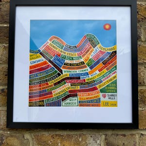 May include: A framed art print featuring a stylised map of Surrey Hills with colourful, segmented sections labelled with various location names like Shere, Box Hill, and Leith Hill, set against a blue sky with a red sun, dated 2026 in the corner.