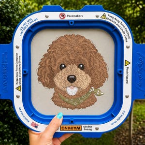 Puede incluir: Un retrato de un perro de pelo rizado marrón bordado sobre tela clara, sujeto en un bastidor de bordado cuadrado azul. El perro lleva un pañuelo verde estampado. El marco incluye advertencias en blanco, como U.S. PAT. NOS. 7,607,399 & 7,918,169.
