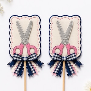 May include: Two rectangular fabric toppers featuring a grey and pink embroidered scissor design on a cream background with scalloped edges. Each topper is decorated with a navy blue gingham bow and layered ribbons in navy, white, and pink on a stick.