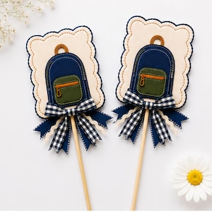 May include: Two decorative toppers featuring a cream-colored scalloped rectangle base with a navy blue backpack applique. Each backpack has an olive green front pocket with a zipper and a brown handle. A navy and white gingham bow sits below.