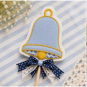 Könnte beinhalten: Ein glockenförmiges Stoffornament in Hellblau mit gelber Stickkante und einem gelben Klöppel in der Mitte. Die Glocke hat ein blaues Blumenmuster im Inneren und ist mit einem dunkelblauen, gepunkteten Band sowie weißer Spitze verziert.