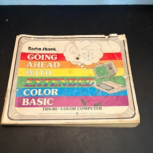 Op de afbeelding: Een vintage Radio Shack handleiding getiteld GOING AHEAD WITH EXTENDED COLOR BASIC voor de TRS-80 COLOR COMPUTER. De kaft heeft een regenboogachtergrond, een cartoon computerfiguur met een gedachtenwolk en witte tekst op gekleurde banen.