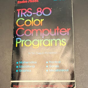 May include: A black paperback book titled Radio Shack TRS-80 Color Computer Programs by Tom Rugg and Phil Feldman. The cover features a rainbow-coloured gradient on the title text and lists categories including Mathematics, Educational, and Games.