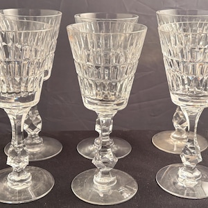 Puede incluir: Un conjunto de cinco copas de cristal transparente con un patrón geométrico facetado en el cáliz y un diseño de tallo multifacetado. Cada copa tiene una base circular ancha y un borde acampanado, mostrando una textura de cristal tallado.