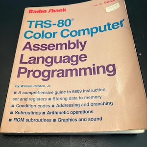 Könnte beinhalten: Ein Taschenbuch mit dem Titel TRS-80 Color Computer Assembly Language Programming von William Barden, Jr. Der Einband ist hellbraun mit rotem, blauem und violettem Text, inklusive Radio Shack Logo und Katalognummer 62-2077.