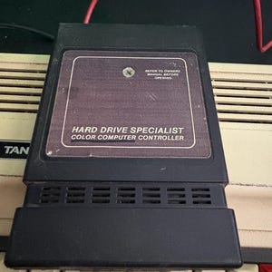 Peut inclure: Une cartouche noire rectangulaire Hard Drive Specialist Color Computer Controller avec une étiquette indiquant "REFER TO OWNERS MANUAL BEFORE OPENING" et "HARD DRIVE SPECIALIST COLOR COMPUTER CONTROLLER" placée sur un clavier d'ordinateur beige.