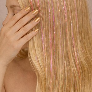 Könnte beinhalten: Blondes Haar mit rosa und schillernden Glitzersträhnen. Die Hand der Person bedeckt ihr Gesicht. Die Nägel sind goldfarben lackiert. Das Haar ist lang und glatt, mit vertikal verlaufenden Glitzersträhnen.