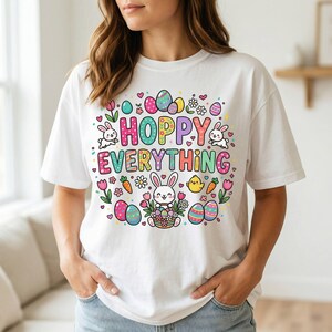 Könnte beinhalten: Weißes T-Shirt mit einem farbenfrohen Ostermotiv. Das Design zeigt den Text "HOPPY EVERYTHING", Ostereier, Hasen, Blumen und Karotten. Ein lässiges, festliches Kleidungsstück.