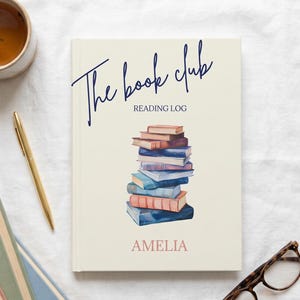 May include: A cream-colored reading log book titled The book club with a stack of watercolor books in the center and the name AMELIA printed in pink at the bottom. The book is surrounded by a mug of tea, a gold pen, and tortoise shell glasses.