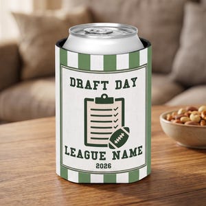 Puede incluir: Un enfriador de latas cilíndrico de espuma con un patrón de rayas verticales en blanco y verde salvia. El centro muestra un gráfico rectangular con el texto DRAFT DAY, LEAGUE NAME y 2026, junto a una ilustración de un portapapeles y un balón de fútbol americano.