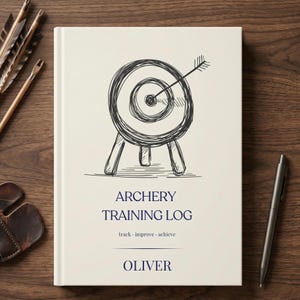 Könnte beinhalten: Ein cremefarbenes Hardcover-Buch mit dem Titel Archery Training Log und einer schwarzen Skizze einer Zielscheibe mit Pfeil auf dem Einband. Der Text lautet Archery Training Log, track improve achieve, und der Name Oliver in Dunkelblau.