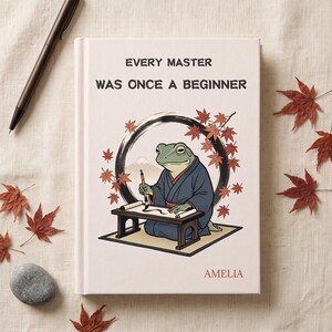 May include: A white hardcover journal featuring the text "EVERY MASTER WAS ONCE A BEGINNER" and "AMELIA" at the bottom. The cover displays a green frog in a blue robe painting on a scroll, framed by a black circle and red maple leaves.