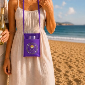 Pode incluir: Um porta-copos de vinho roxo com uma alça de ombro, segurando uma taça de vinho transparente. O porta-copos tem um design floral e o texto "SOL&WINE". O fundo mostra uma praia e o oceano.