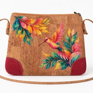 Puede incluir: Un bolso bandolera de corcho con un estampado de acuarela vibrante de un colibrí y hojas tropicales. El bolso tiene un cierre de cremallera superior, una correa de hombro fina de corcho y detalles de ante rojo oscuro en las esquinas inferiores.