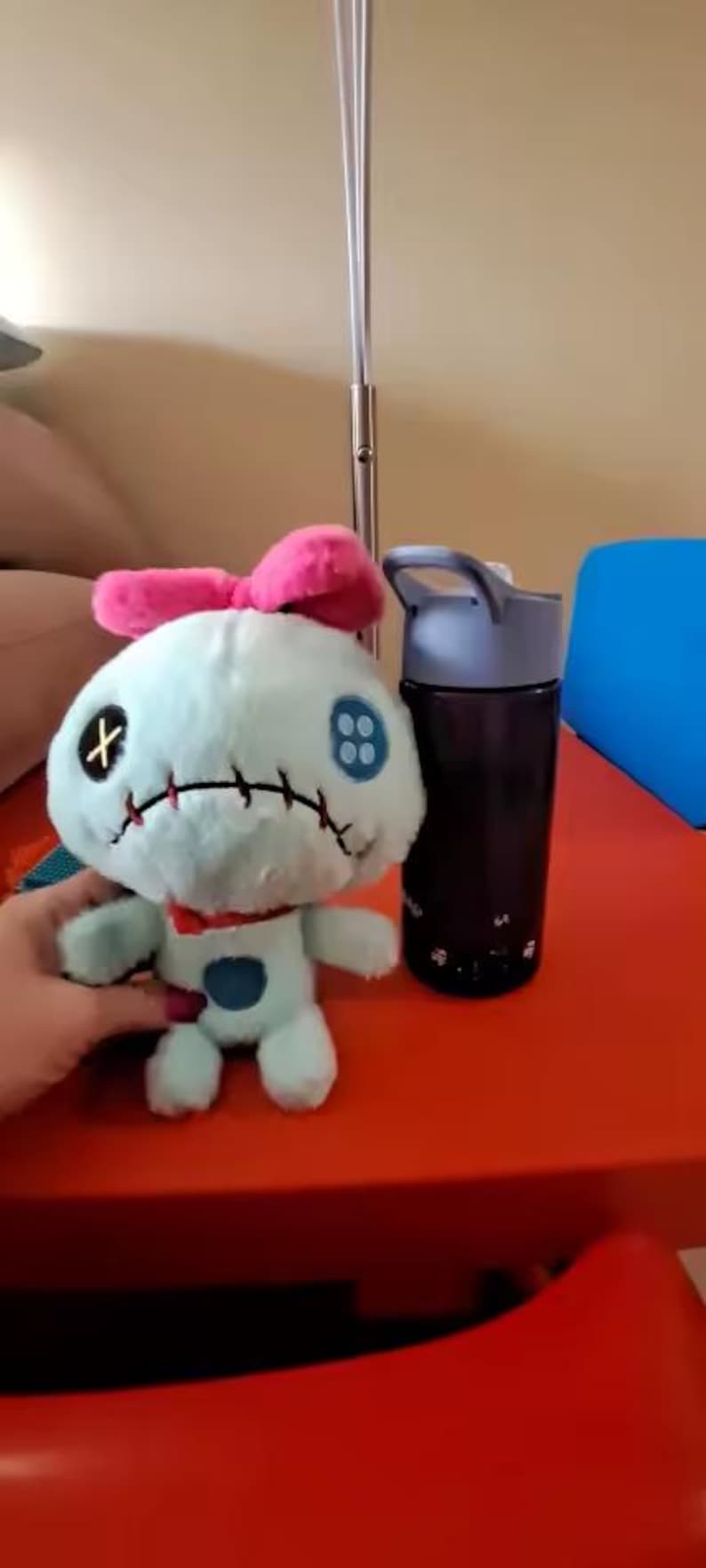 May include: A plush toy of a blue alien-like creature with a pink bow, black stitched features, and a red collar. A black water bottle with a gray lid sits next to the toy on a red surface.
