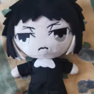 May include: A plush doll with a black hairstyle and a black outfit. The doll has a white face with black eyes and a small, downturned mouth. The doll's outfit includes a white collar.