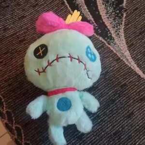 May include: A plush doll in the shape of a cartoon character. The doll is light blue with a pink bow, red collar, and yellow accents. It has stitched facial features, including an X for an eye and a stitched mouth.
