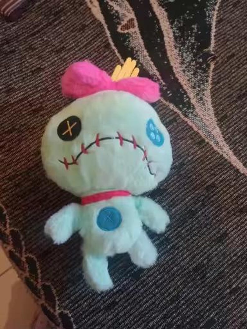 May include: A plush doll in the shape of a cartoon character. The doll is light blue with a pink bow, red collar, and yellow accents. It has stitched facial features, including an X for an eye and a stitched mouth.