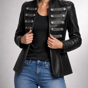 Puede incluir: Una chaqueta de cuero negro con cuello alto, mangas largas y un cierre frontal adornado con botones metálicos de tono plateado y detalles decorativos de cordón trenzado. La chaqueta incluye charreteras en los hombros con botones plateados a juego.