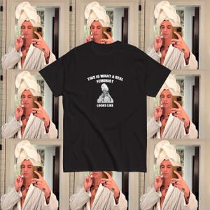 Peut inclure: Un t-shirt noir avec une impression graphique blanche représentant une personne portant une serviette blanche sur la tête et un peignoir blanc. Le texte au-dessus et en dessous du graphique indique, This is what a real feminist looks like.