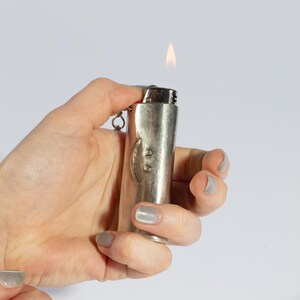 Op de afbeelding: Een zilverkleurige aansteker met een vlam aan de bovenkant. De aansteker heeft een gebogen ontwerp aan de voorkant. Een kleine ketting is aan de onderkant bevestigd. De aansteker wordt in een hand gehouden tegen een witte achtergrond.