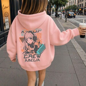 May include: A light pink hoodie with a graphic on the back featuring an anime-style character wearing headphones. The graphic includes the text "K-POP", Japanese characters, and "CREW AMELIA". The hoodie has a hood and long sleeves.