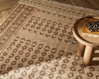May include: A rectangular area rug featuring a beige and tan base with a repeating dark brown floral pattern and a fringed edge. A round, light brown side table sits on the rug, holding a yellow book topped with a cluster of dark brown seeds.