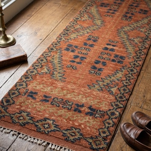 Könnte beinhalten: Ein orangefarbener Läufer mit einem geometrischen Muster in Marineblau und Olivgrün. Der Teppich hat einen Fransenrand und liegt auf einem Holzboden. Braune Leder-Loafers sind sichtbar. Der Teppich misst etwa 200 cm in der Länge.