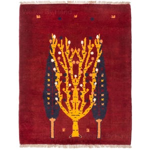 Puede incluir: Una alfombra rectangular con fondo rojo intenso que presenta un motivo de árbol central amarillo flanqueado por dos árboles oscuros tipo ciprés. El diseño incluye pequeños detalles en naranja y blanco, con el texto Weave By Hand visible.