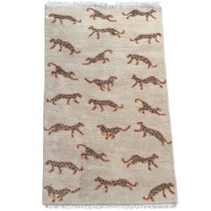 May include: A rectangular area rug featuring a light beige background with a repeating pattern of small, dark brown and grey spotted feline figures in various running poses. The rug has a short pile texture and white fringe along both ends.
