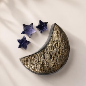 Pode incluir: Um objeto em forma de meia-lua com uma superfície frontal texturizada de cor dourada e bordas escuras, acompanhado por três pequenas peças translúcidas em forma de estrela de cor roxo profundo com manchas escuras perto da lua.