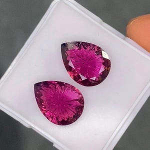 May include: Two pear-shaped, faceted gemstones with a vibrant magenta-pink colour. Each stone features a distinct radial, star-like pattern carved into the surface, creating a textured appearance within the translucent, deep pink material.