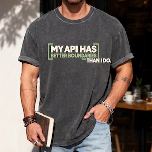 May include: A charcoal gray short-sleeve t-shirt with a crew neckline and rolled sleeves. The chest features the text "MY API HAS BETTER BOUNDARIES THAN I DO." printed in white and light green, enclosed in a rectangular bracket design.