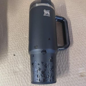 Op de afbeelding: Een donkerblauwe Stanley reismok met een handvat en een bloemenontwerp ge&euml;tst op de bodem. Het Stanley-logo is zichtbaar. De mok heeft een zwart deksel en is ontworpen om dranken warm of koud te houden.